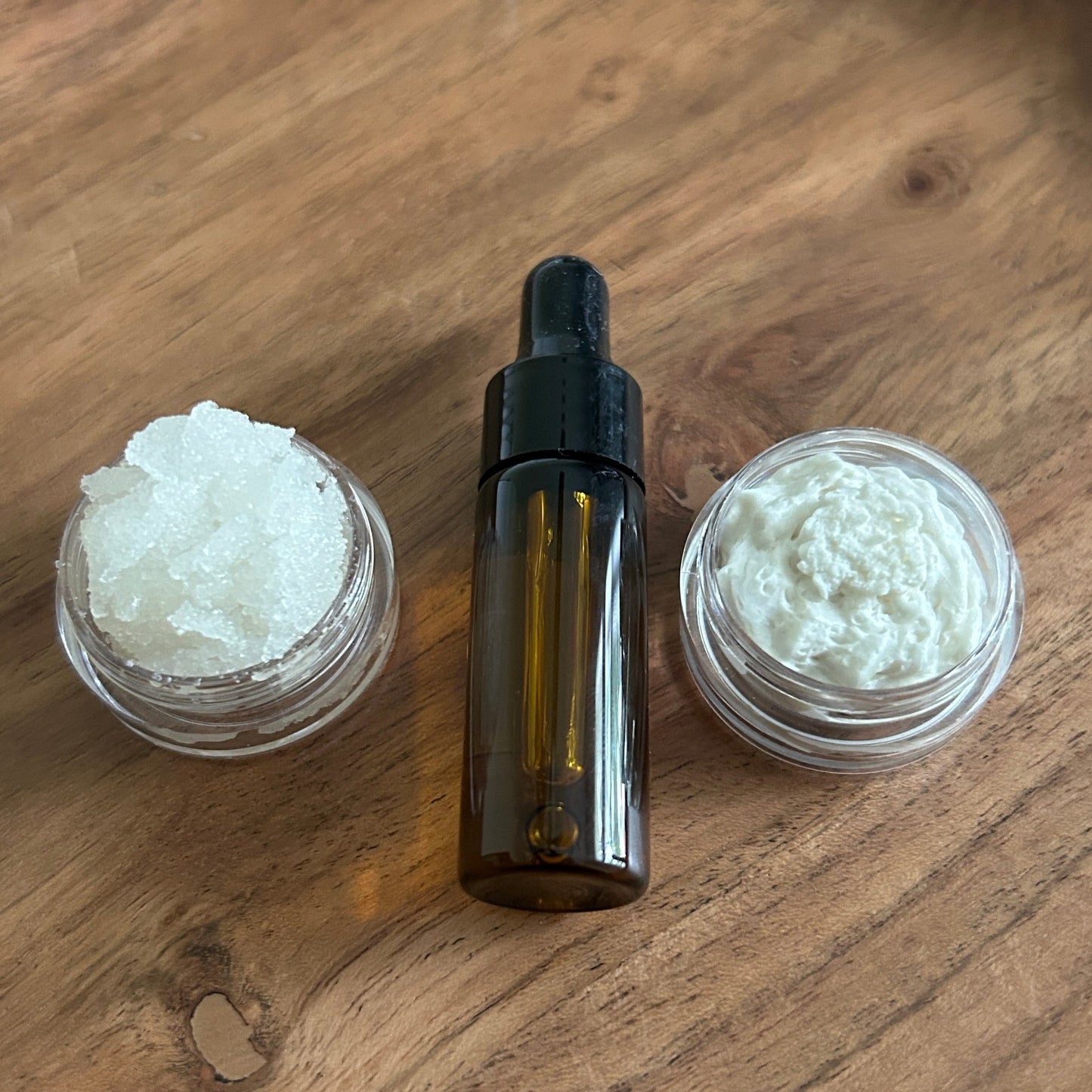 one small container with body scrub, one amber glass dropper bottle of oil, and one small container with body butter on a wooden surface