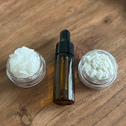 one small container with body scrub, one amber glass dropper bottle of oil, and one small container with body butter on a wooden surface